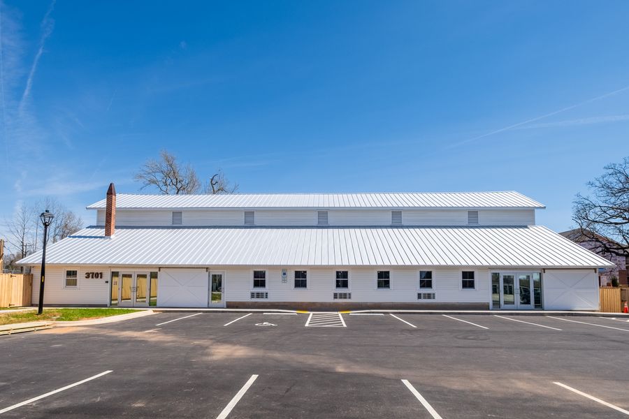 The school building by day: a long white timber-framed barn with a standing-seam metal roof, clerestory vents and a brick chimney, viewed straight on from the parking lot.