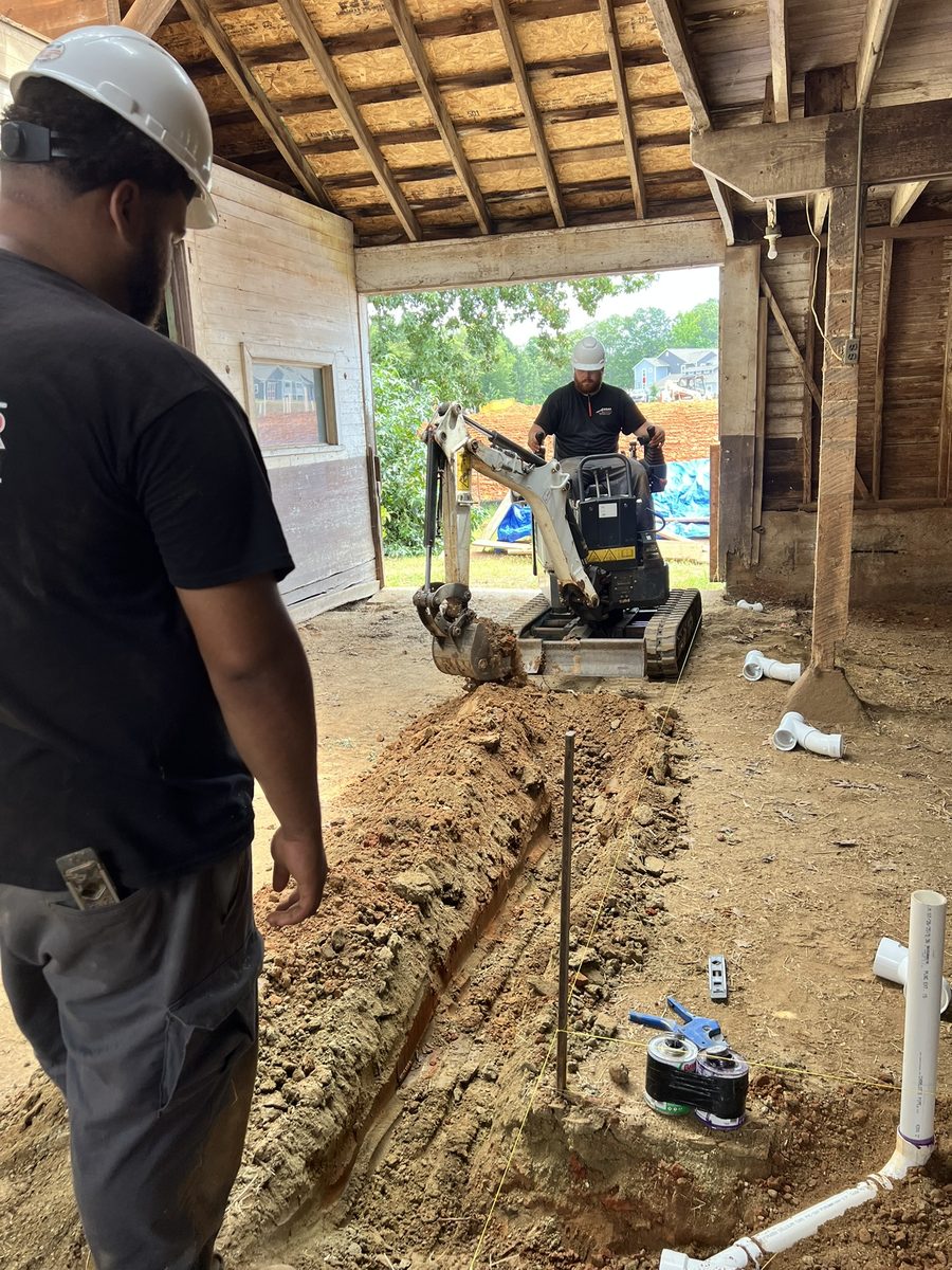 Inside the barn during construction: a compact excavator digs a service trench through the dirt floor while two workers in hard hats look on, with white PVC drainage fittings laid out ready on the ground.