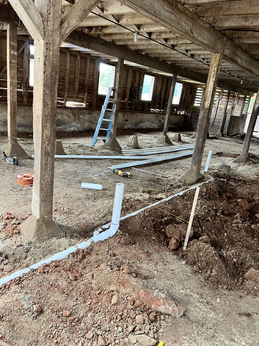 Plumbing rough-in inside the barn: white PVC drainage runs laid along string lines through open trenches, between the original timber posts on their concrete footings.