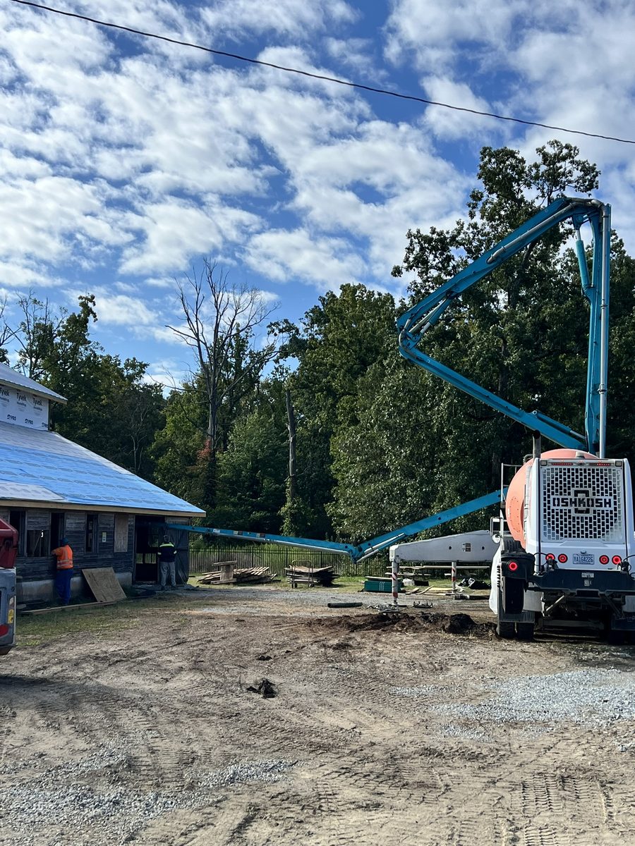 A concrete pump truck parked beside the barn with its boom extended over the building, which is wrapped in blue roof underlayment, as workers in hi-vis stand at an open doorway.