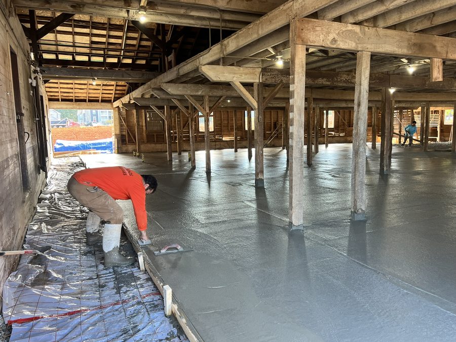 A concrete slab being poured inside the barn: a worker in an orange shirt kneels to trowel the wet edge while another screeds in the background, the fresh concrete flowing around each original timber post.