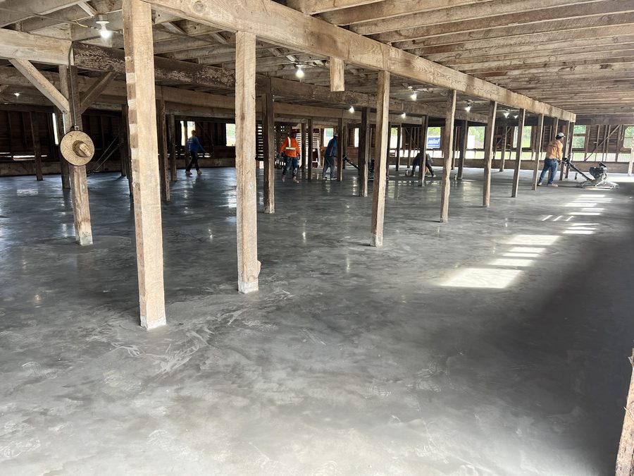 The finished slab inside the barn, wet and reflective, with a crew of workers power-trowelling the surface between rows of original timber posts.