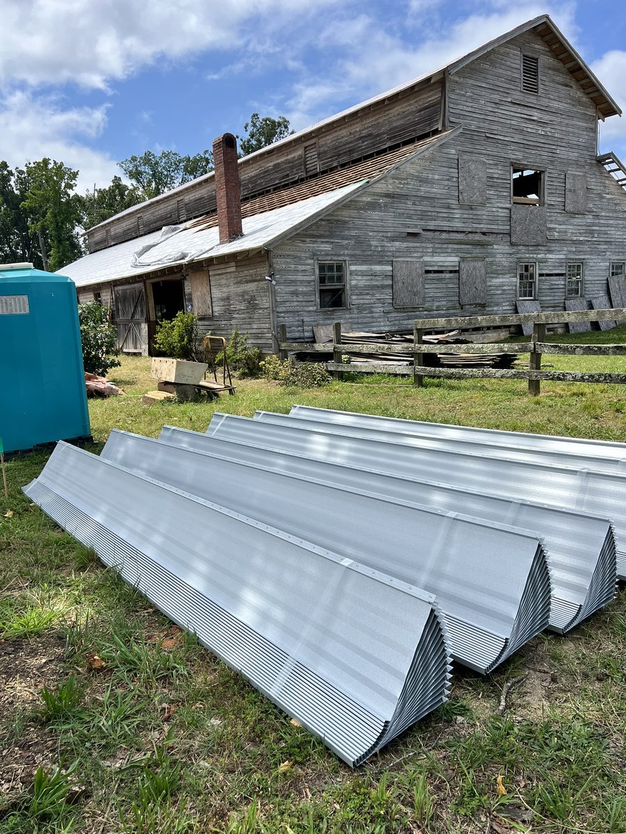 Stacks of new standing-seam metal roof panels laid out on the grass in front of the barn, which still carries its weathered grey siding and part of its old roof.