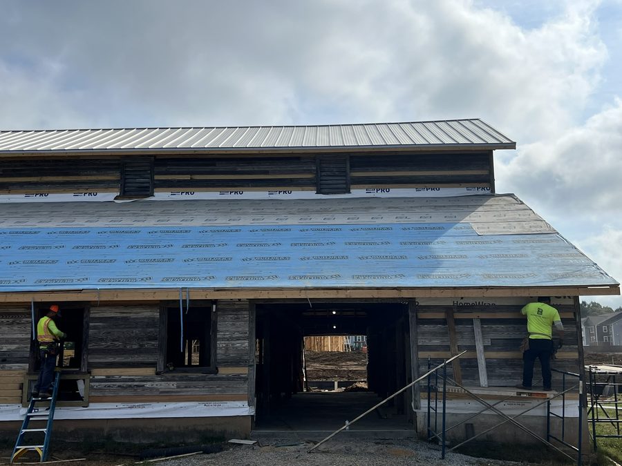 The barn part-way through re-roofing: house wrap across the lower walls, new timber trim, and fresh metal roofing being laid over the underlayment while a worker moves along the eaves.