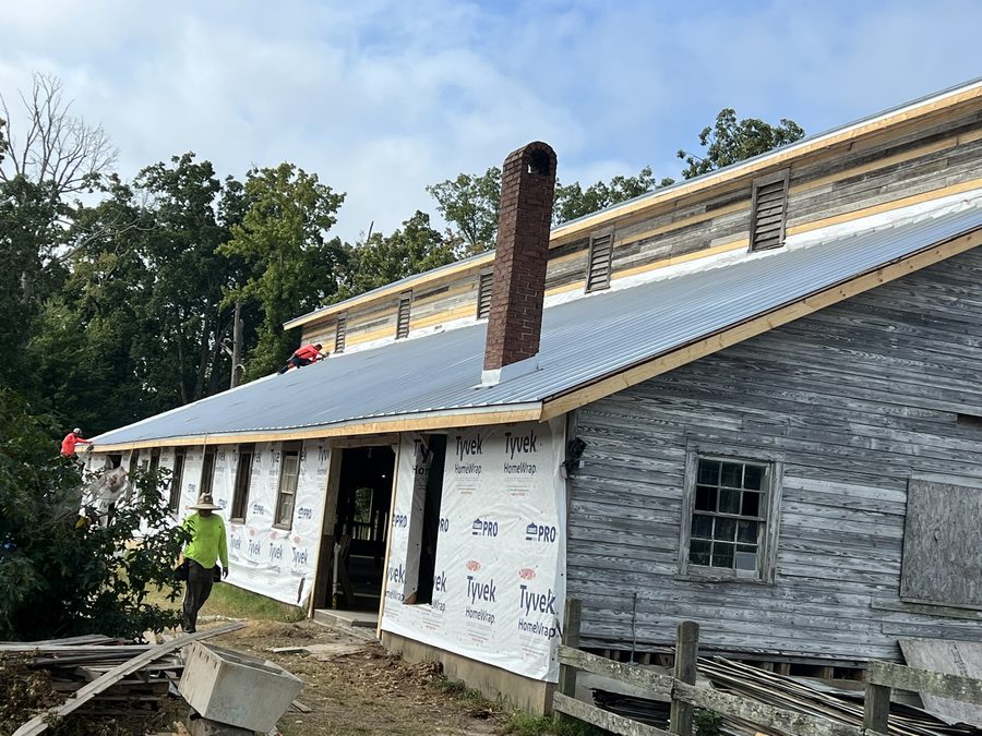Two workers on ladders and scaffold fitting house wrap and roof underlayment to the gable end of the barn, the original weathered boards still visible between the new material.