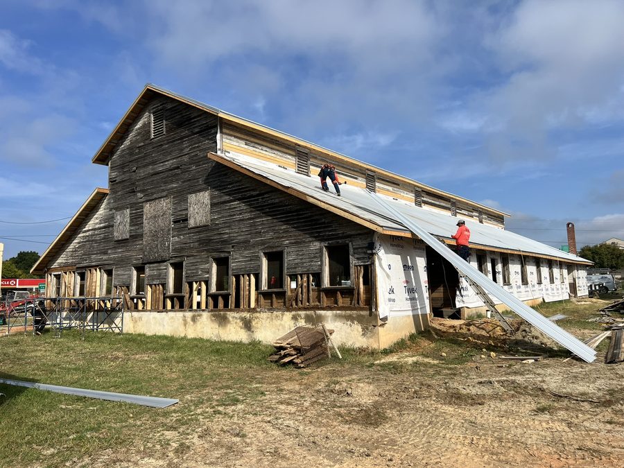 A wide view of the barn with roofing nearly complete: new standing-seam panels along the lower roof, house wrap on the walls, a worker carrying a panel up the slope, and the original grey siding still in place above.