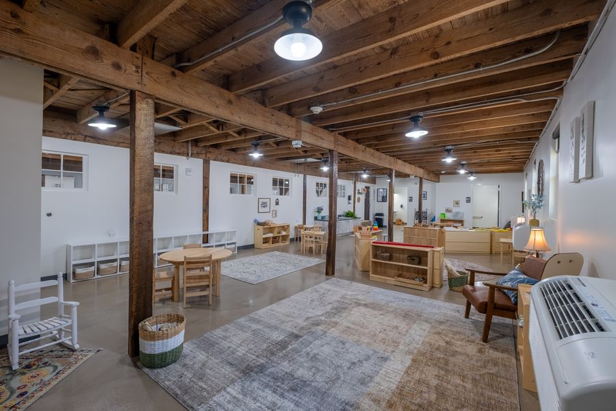A wide classroom under exposed timber beams and posts, with low wooden shelves, a round child-sized table, woven baskets and a white rocking chair on a soft rug.