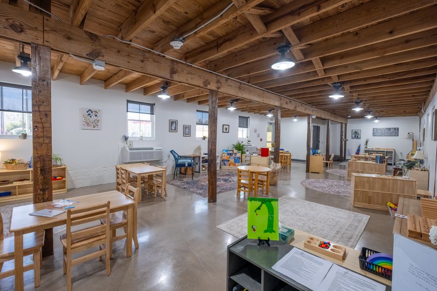 A Giving Tree classroom with low wooden shelves of Montessori materials, child-sized tables and work rugs beneath exposed timber beams.