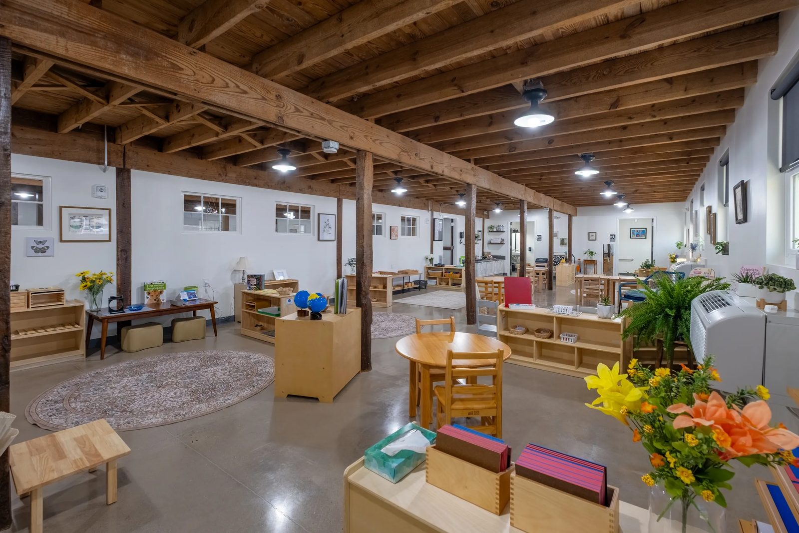A Giving Tree classroom under exposed timber beams and posts, with low wooden shelves of Montessori materials, child-sized tables, work rugs and plants on polished concrete floors.
