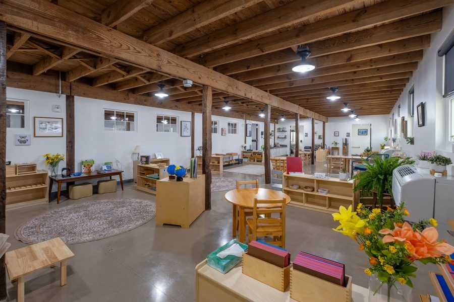 A wide view down a classroom: timber posts and beams overhead, round rugs, low shelves of materials, plants on the windowsills and framed prints on the walls.