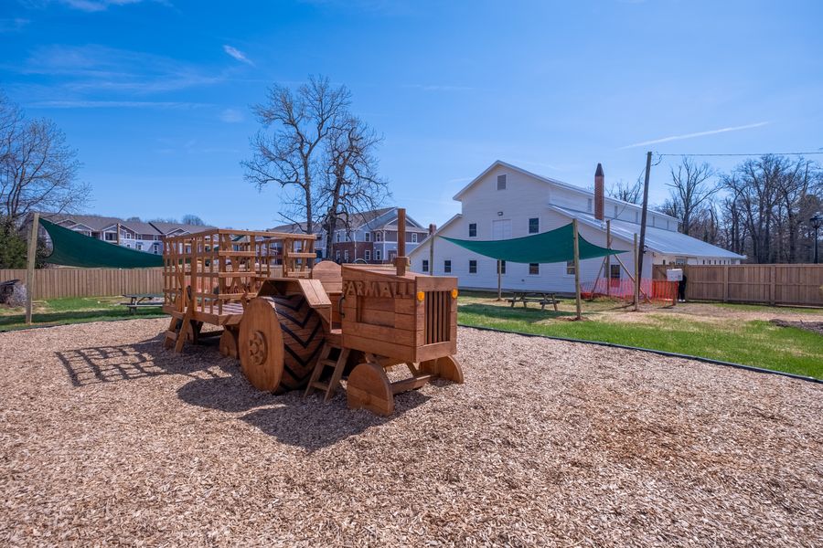 A fenced play yard with a large wooden tractor and wagon climbing structure on wood chips, green shade sails overhead and the school building behind.