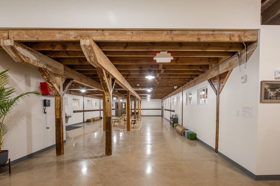 A long open common room under exposed timber beams and posts, with white walls, a row of coat hooks, a drinking fountain, low stools and tables on a rug.
