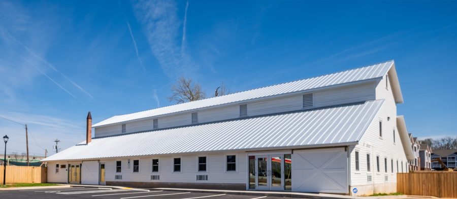A long white timber-framed barn with a standing-seam metal roof, clerestory vents and a brick chimney, seen across the striped parking lot under a clear sky.