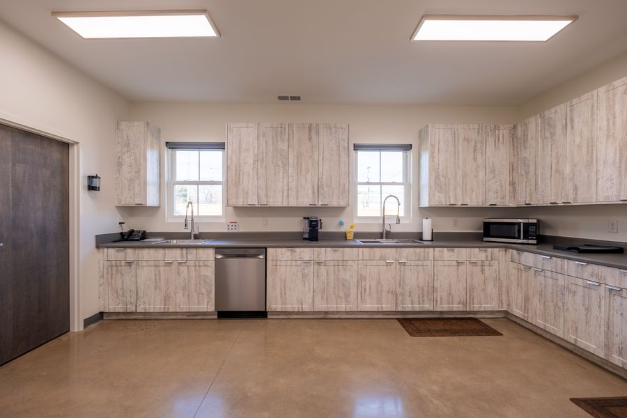 A commercial-style kitchen with pale wood-effect cabinetry, grey countertops, two sinks, a dishwasher and microwave, lit by windows and ceiling panels.