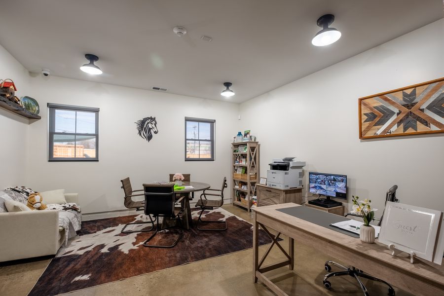 A front office with white walls, a wooden desk, a round meeting table with chairs on a hide rug, a bookshelf of family resources, a printer and a monitor showing camera views.