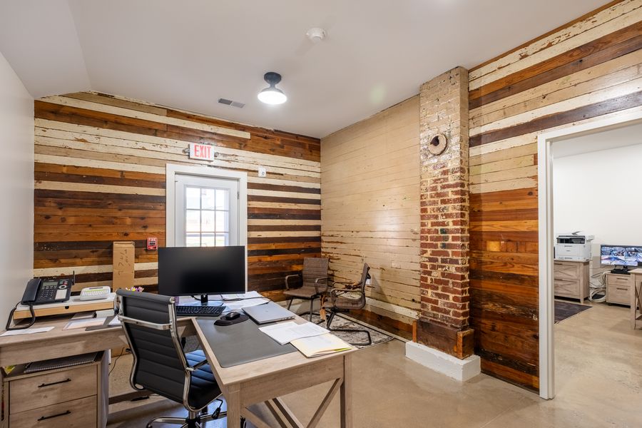 An office with walls clad in reclaimed boards in mixed tones and an exposed brick chimney, a desk with a monitor and a phone, and chairs for visitors.