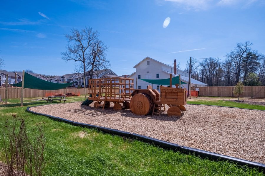 A large wooden play tractor and trailer with a slide, set on wood-chip surfacing in the fenced yard, with green shade sails and the school building behind.