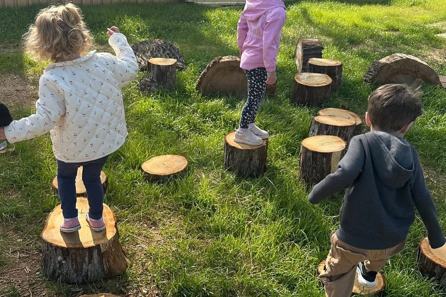 Three children stepping between cut tree rounds set into the grass, arms out for balance.