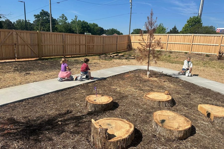 Children riding small balance bikes along a concrete path through the yard, with cut tree rounds and a young tree nearby.