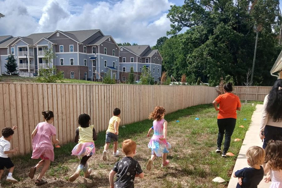 Children running across the grass beside the fence line, with a teacher walking ahead and cones set out on the ground.