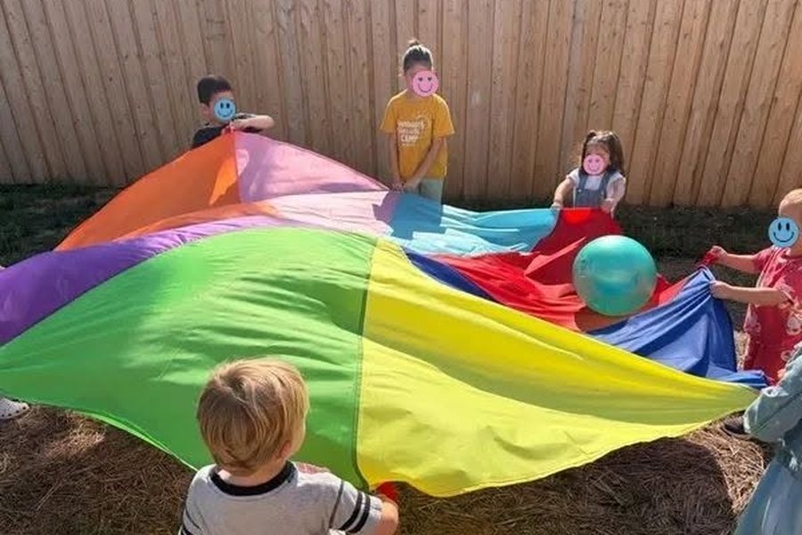 Children standing around a large rainbow parachute in the fenced yard, lifting it together with a ball resting on top.