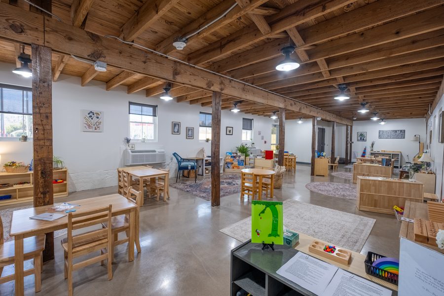 The Primary classroom with work tables on rugs, low shelves of materials, and a copy of The Giving Tree standing on a shelf in the foreground.