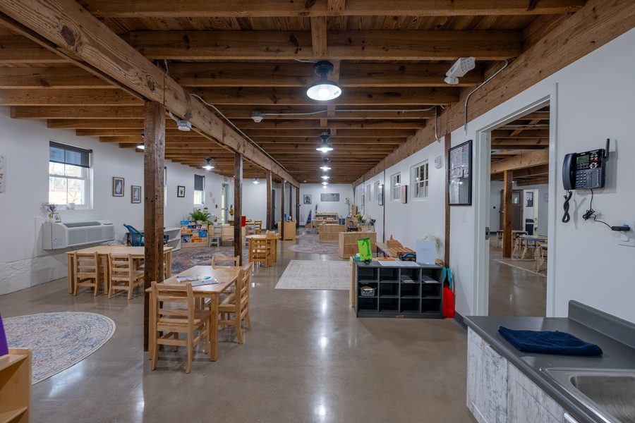 A long view down the Primary classroom with cubby storage, work tables, a wall telephone and a doorway through to the adjoining room.