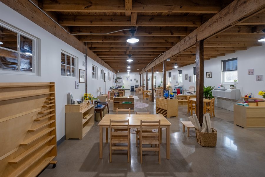 The Primary classroom with a tall rolling shelf rack on the left, work tables on rugs, and globes and an atlas on a low unit.