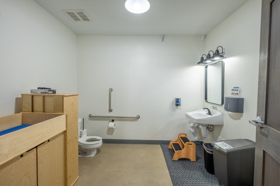 A children's restroom with a low toilet, grab bars, a wooden changing table, a step stool at the sink and a paper towel dispenser within reach.