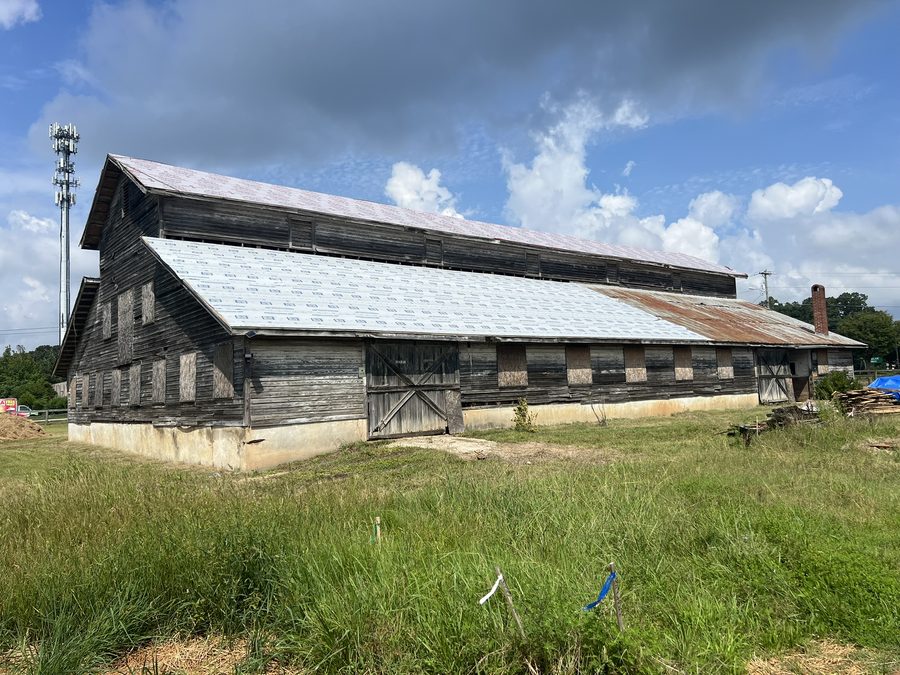 The barn before restoration: weathered grey board siding, boarded-up window openings, a rusted metal roof partly stripped and re-covered with new underlayment, standing in long uncut grass with survey stakes in the foreground.