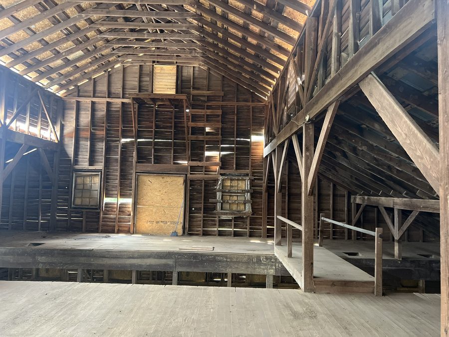 A stall bay with slatted board partitions, a ventilation fan set into the framing, and straw across the dirt floor.