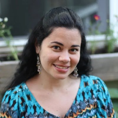 Hanan Salim, Director — smiling woman with dark hair and blue patterned dress outdoors.