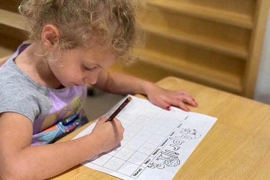 A child seated at a table, pencil in hand, working carefully through a printed letter-formation sheet.