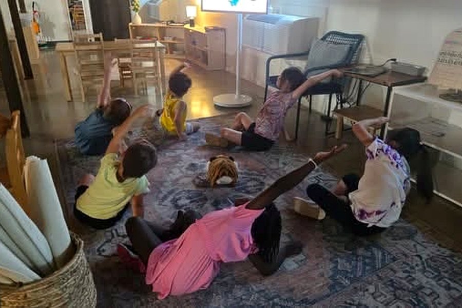 Children lying on a patterned rug in the classroom with arms stretched overhead during a movement session.