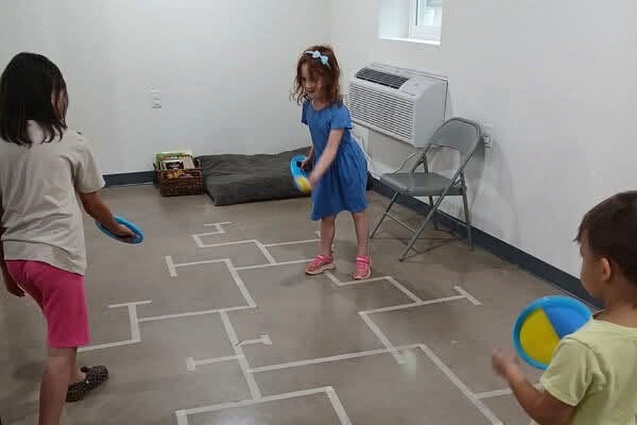 Three children playing a bean-bag game on a floor course marked out with tape in the common room.