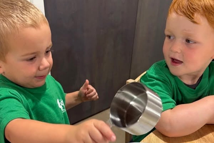 Two children in green school shirts at a table, one spooning butter onto a baked potato on a plate.