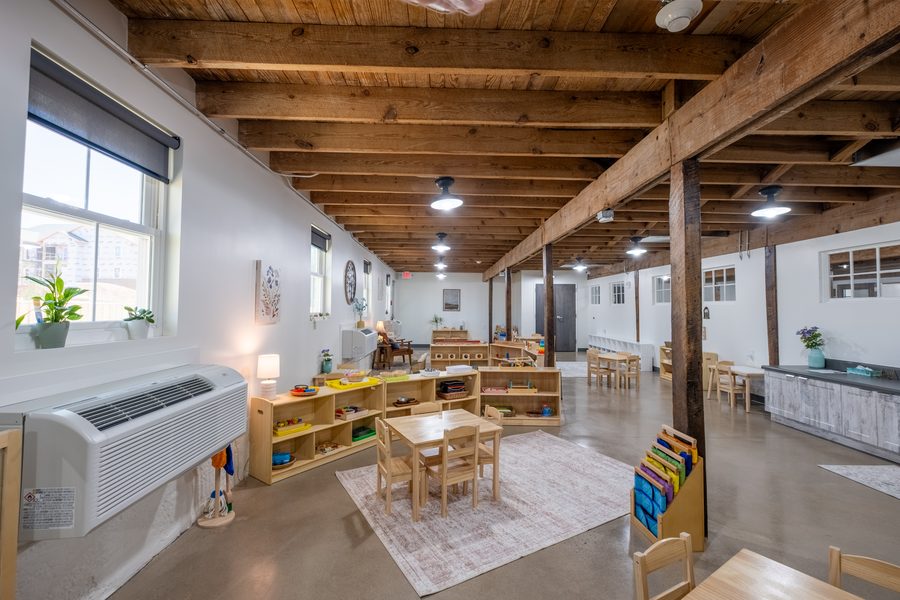 The Toddler classroom from the doorway: tall windows with plants on the sill, low open shelving with trays of work, and a table and chairs on a soft rug.