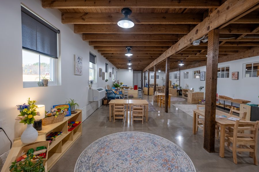 A long view down the Toddler classroom past timber posts, with a round rug in the foreground, tables and shelves of materials along the wall.